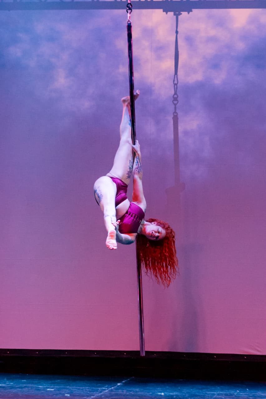 Paola Liébana invertida y agrupada sobre el caño suspendido, en una presentación de pole flying.